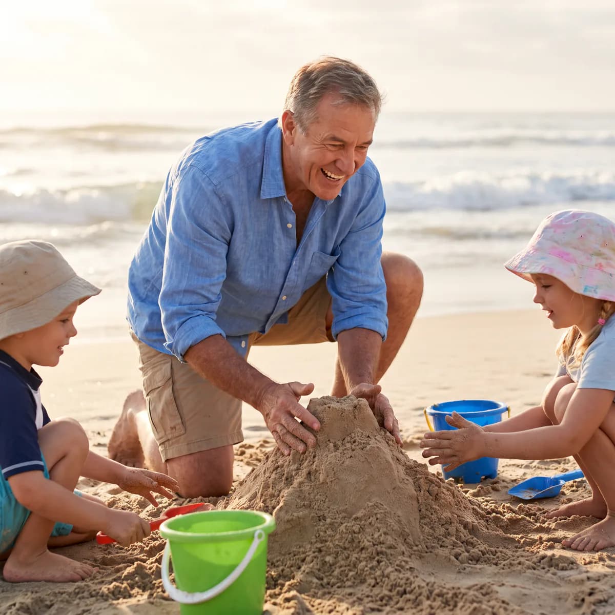 A gray-haired grandfather kneels in the sand building a sandcastle with three young grandchildren in sun hats, ocean sunset behind them